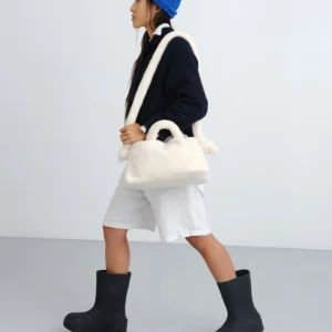 A person in a blue knitted hat, black jumper, white shorts, and black boots walks across a light grey Estonian-inspired room, carrying the Mini Ona Sherpa Bag - Sand over one shoulder and holding it by the handle. Gate21.ee
