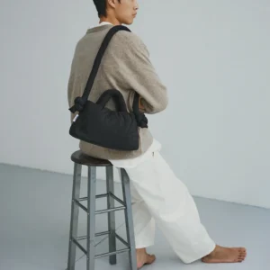 A person with short dark hair sits barefoot on a metal stool, facing sideways. Wearing a beige jumper, white trousers, and carrying the Mini Ona Soft Bag - Black, they offer chic inspiration for unique shopping ideas in Tallinn. Background is plain and light-coloured. Gate21.ee
