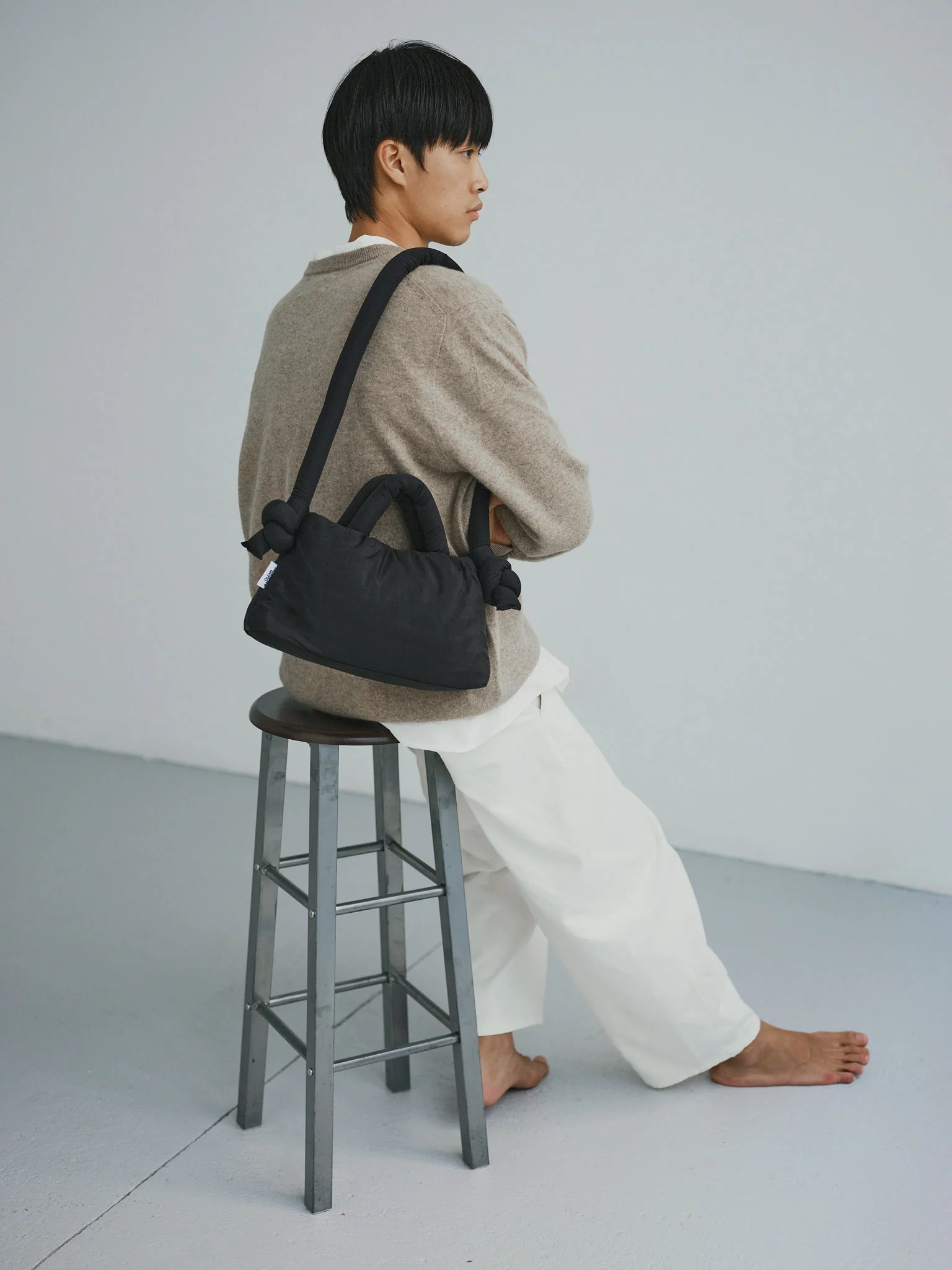 A person with short dark hair sits barefoot on a metal stool, facing sideways. Wearing a beige jumper, white trousers, and carrying the Mini Ona Soft Bag - Black, they offer chic inspiration for unique shopping ideas in Tallinn. Background is plain and light-coloured. Gate21.ee