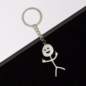 The Zen - Keyring Man - KÕIK is a metal keyring featuring a smiling stick figure holding up both middle fingers, set against a diagonally split black and white background—a bold Estonian design available in Tallinn concept shops. Gate21.ee