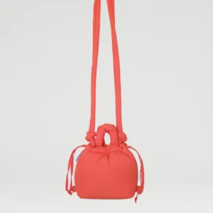 The Micro Ona Soft Bag - Coral is a compact, vibrant red fabric bag with a drawstring fastening, long shoulder straps, and a knotted handle, displayed against a plain white background—an eye-catching gift shop piece. Gate21.ee
