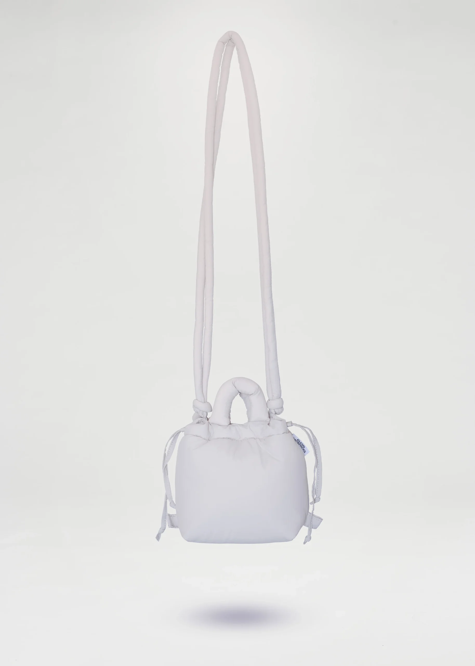 The Micro Ona Soft Bag - Sand is a small, sand-coloured, square handbag with a drawstring fastening, short handles, and a long padded shoulder strap, showcasing Estonian design, photographed on a plain white background. Gate21.ee
