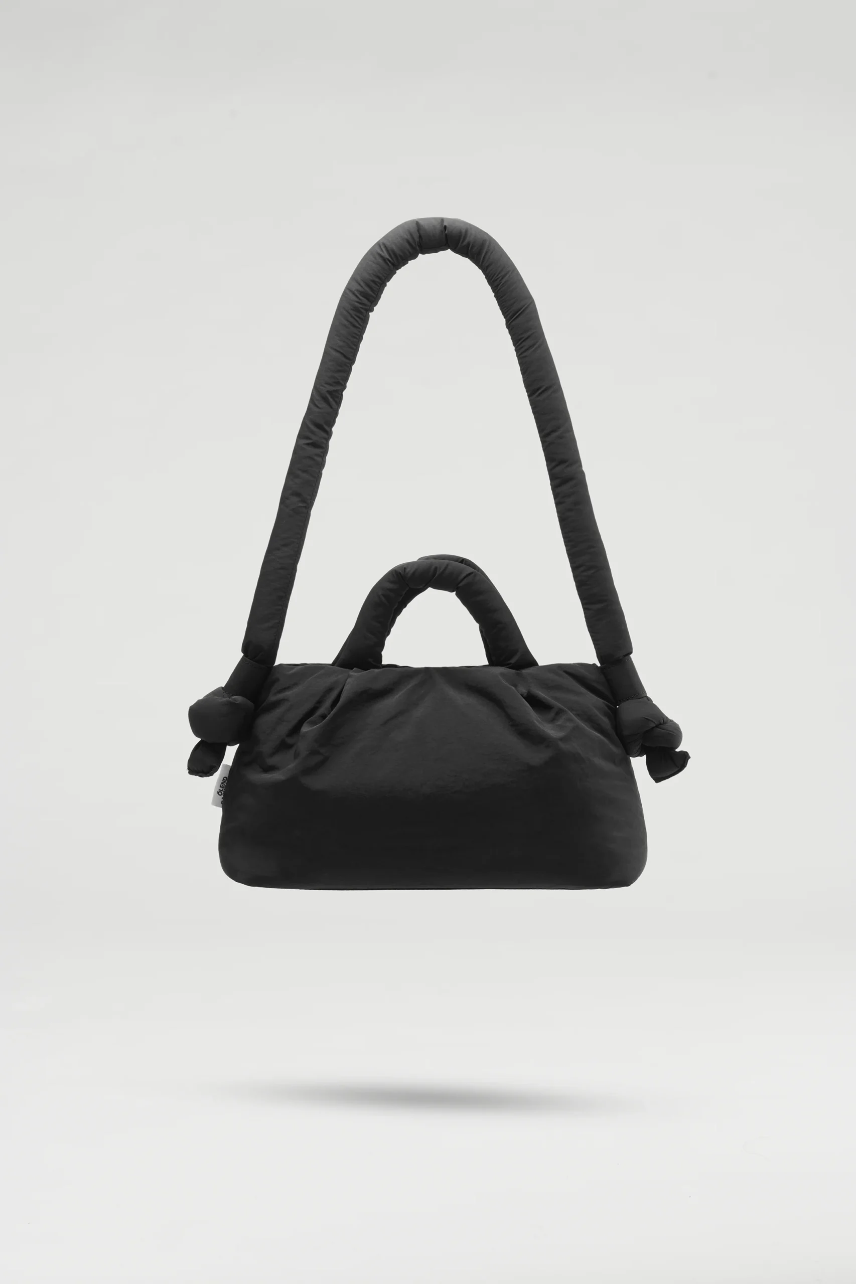 The Mini Ona Soft Bag - Black, featuring a large curved shoulder strap and two small handles, is displayed suspended against a plain light grey background—an elegant addition to any unique gift shop. Gate21.ee