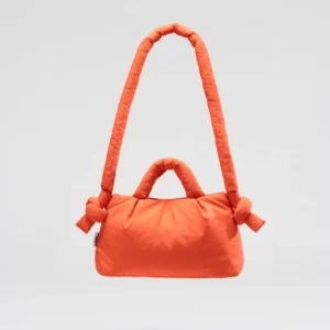 The Mini Ona Soft Bag - Coral is a padded orange handbag with knotted details on its long shoulder strap and two short handles, shown against a light grey background—ideal for curated accessory lovers seeking unique concept shop finds. Gate21.ee
