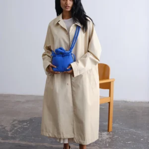 A woman in a long beige coat and beige shoes stands indoors, holding the Micro Ona Soft Bag in Cobalt Blue from a curated Estonian design gift shop. A wooden chair is behind her on the concrete floor. Gate21.ee