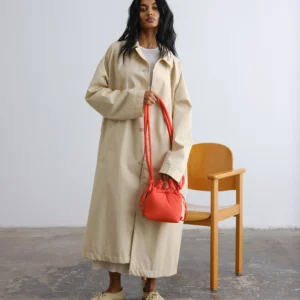 A person with long dark hair stands indoors wearing a long beige trench coat and beige shoes, holding the Micro Ona Soft Bag - Coral—perfect for Tallinn shopping—with a wooden chair behind them. Gate21.ee
