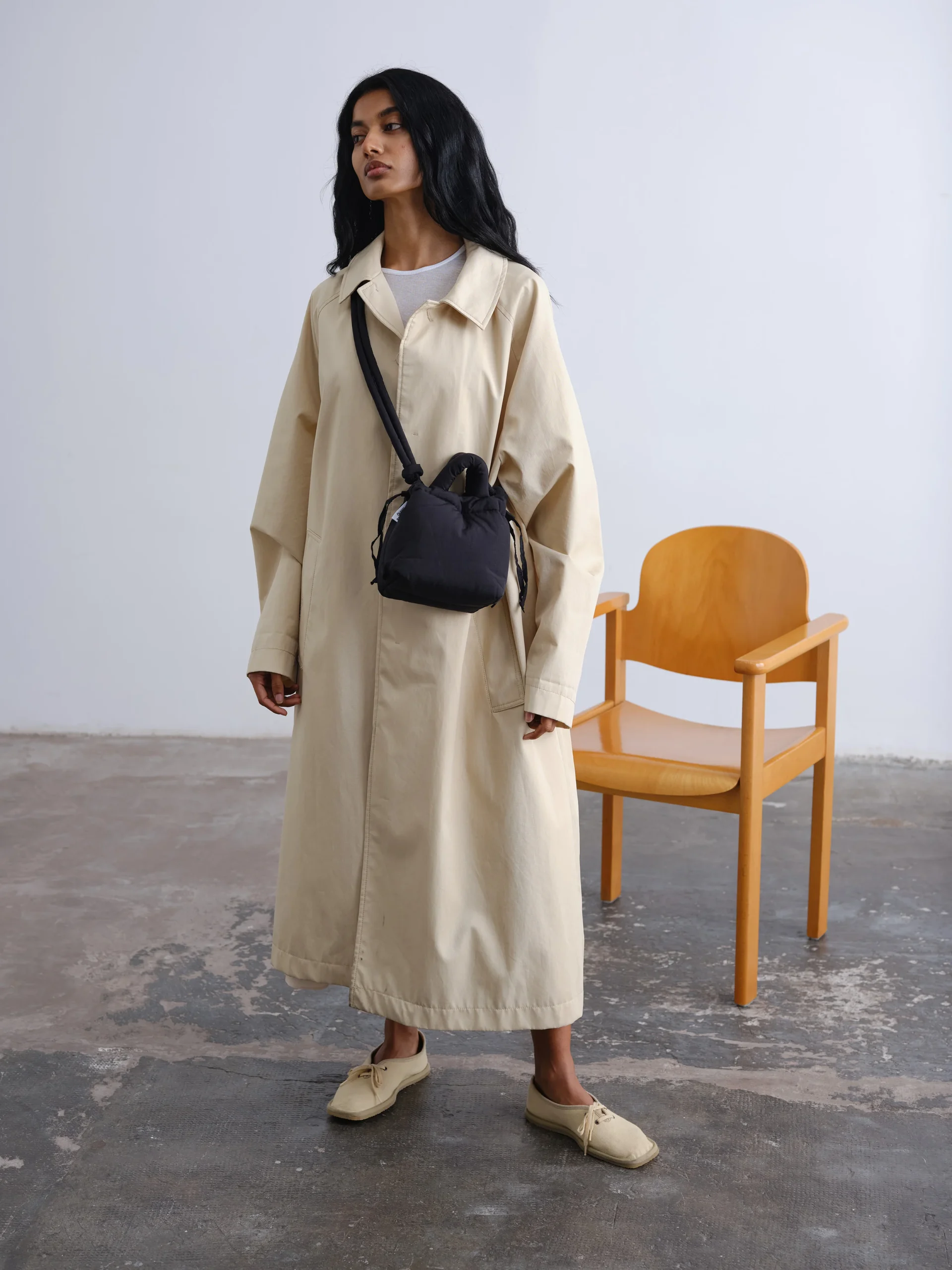 A person with long dark hair stands indoors wearing a beige trench coat, light shoes, and the Micro Ona Soft Bag - Black, creating an effortlessly curated look. A wooden chair rests against the white wall in the background. Gate21.ee