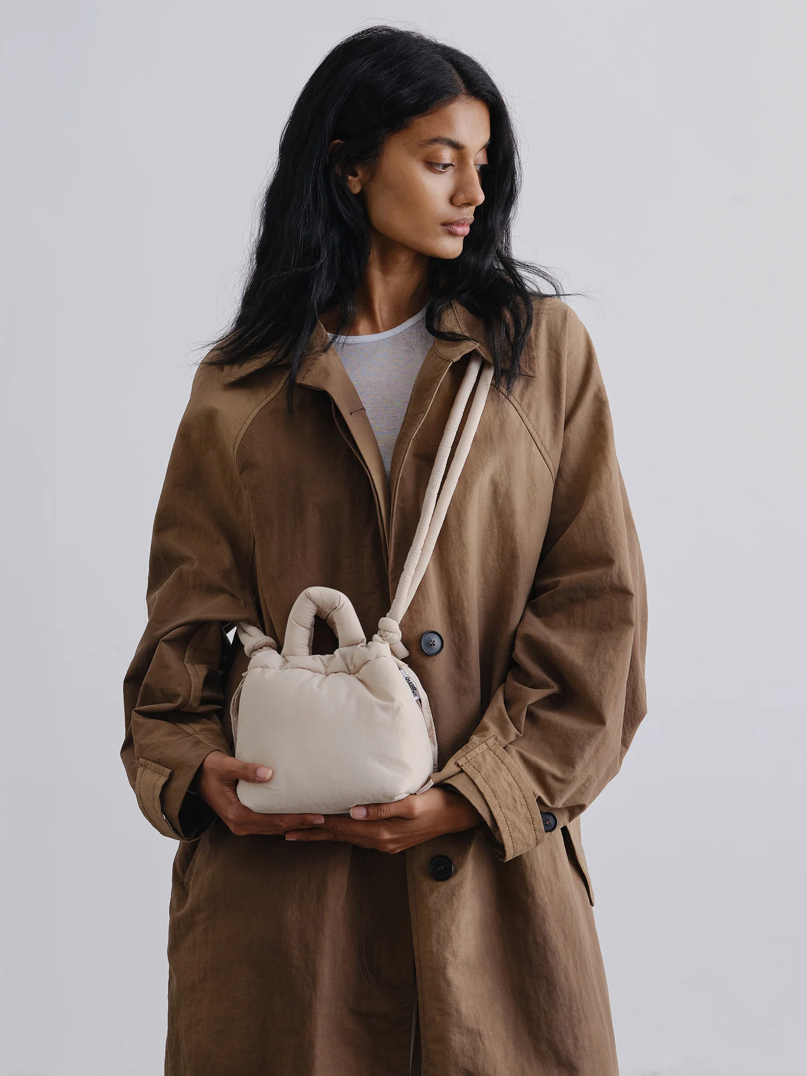 A person with long dark hair, in a brown trench coat over a light shirt, looks to the side while holding the Micro Ona Soft Bag - Sand—an elegant, off-white padded handbag—against a plain light background. Gate21.ee