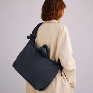 A person with shoulder-length brown hair in a light beige coat faces away from the camera, carrying the Ona Soft Bag - Coal, a large, padded tote inspired by Estonian design, over one shoulder against a light plain background. Gate21.ee