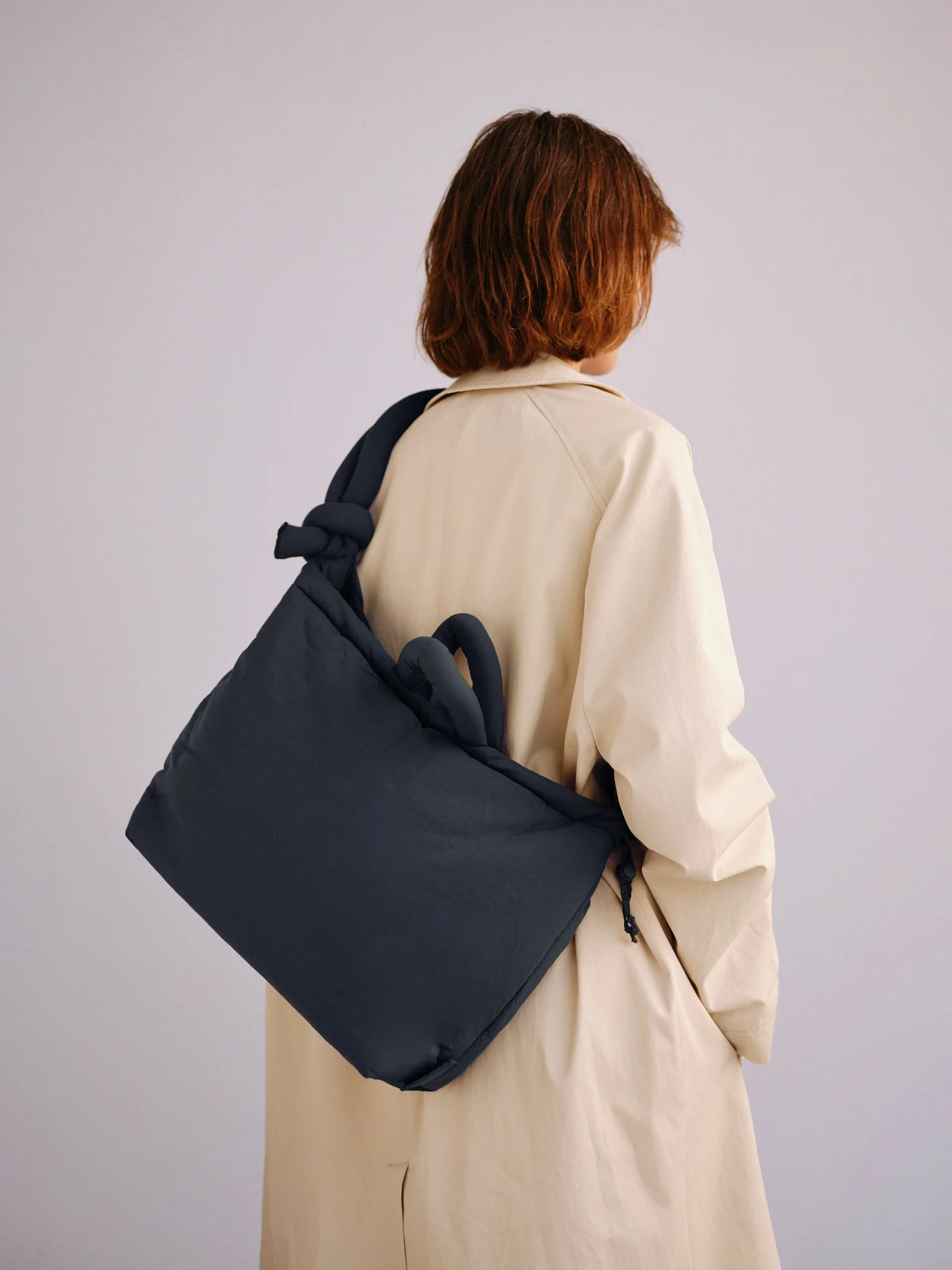 A person with shoulder-length brown hair in a light beige coat faces away from the camera, carrying the Ona Soft Bag - Coal, a large, padded tote inspired by Estonian design, over one shoulder against a light plain background. Gate21.ee