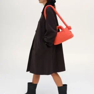 A person in a long dark coat and black boots walks sideways, carrying the Mini Ona Soft Bag - Coral over their shoulder against a plain white background. Gate21.ee