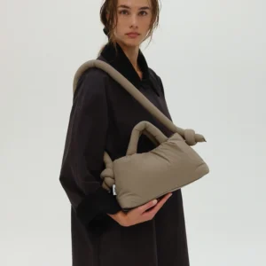 A person in a loose dark coat stands against a plain light background, holding the Mini Ona Soft Bag in Sage—ideal for those who love curated concept shop finds and Tallinn shopping. Gate21.ee