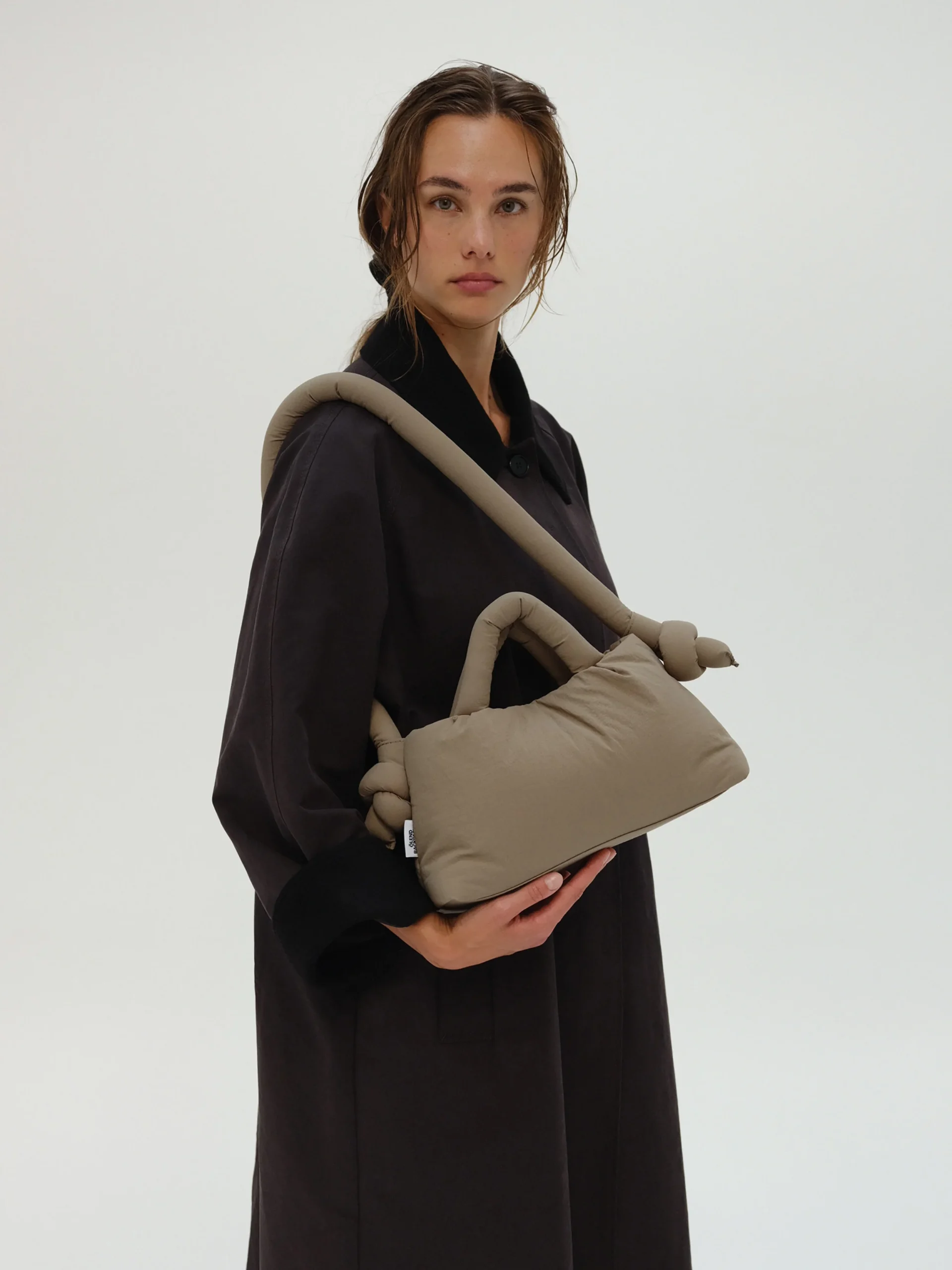 A person in a loose dark coat stands against a plain light background, holding the Mini Ona Soft Bag in Sage—ideal for those who love curated concept shop finds and Tallinn shopping. Gate21.ee