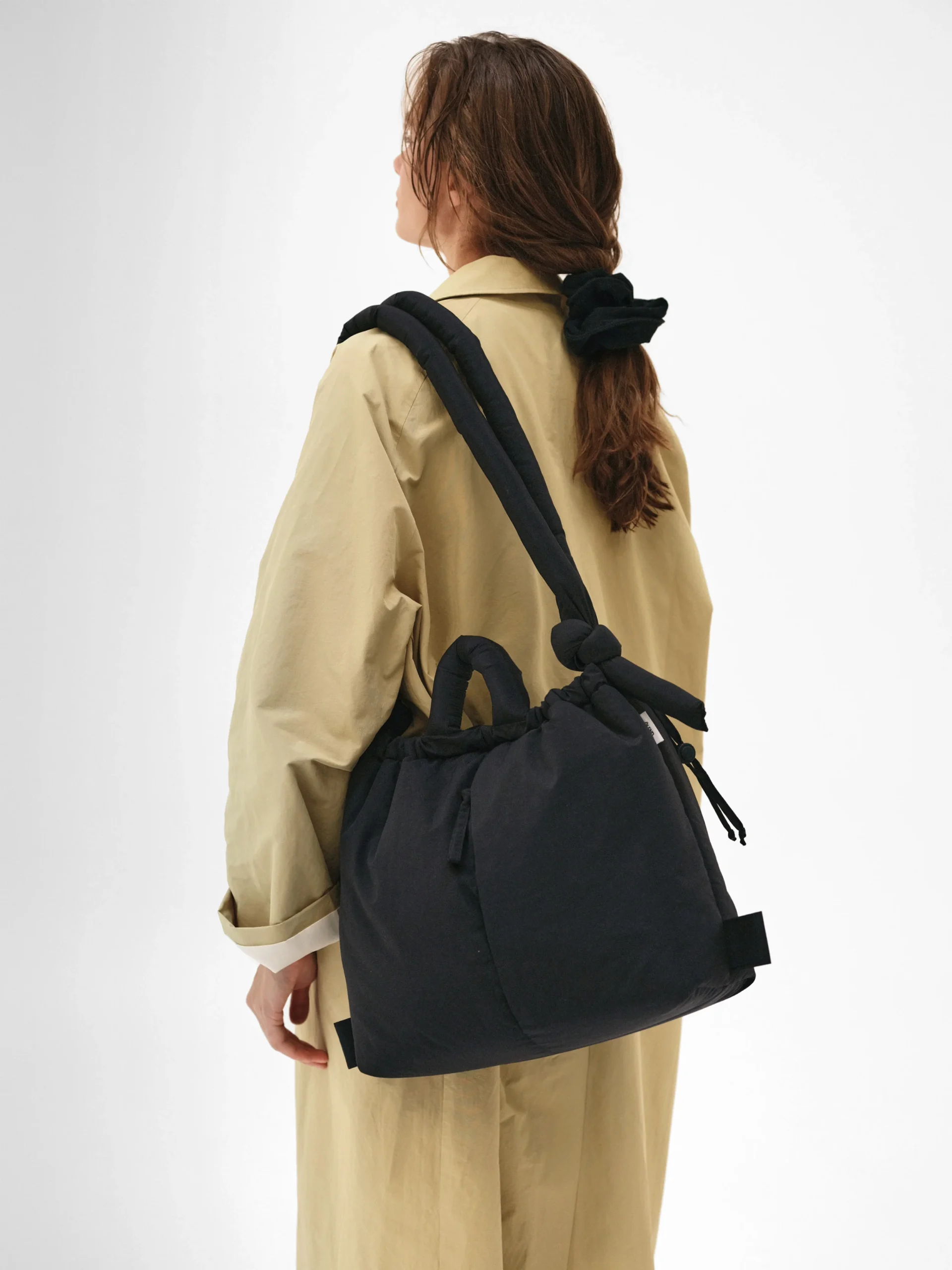 A person with long brown hair, in a beige coat and black hair tie, carries the Ona Soft Bag - Black—an Estonian-designed accessory that stands out for those seeking unique finds when shopping in Tallinn. Gate21.ee