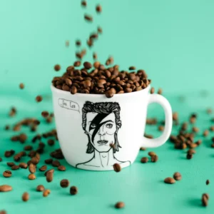 The Glam Rocker - Big cup is a white mug with a black lightning bolt face paint design, showcasing Estonian style; it’s shown filled and surrounded by coffee beans on a teal background. Gate21.ee