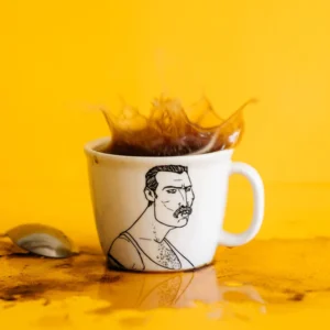 The Mercurial Champion - Large cup, a white mug with a black moustached man line drawing and Estonian design flair, is shown on a yellow surface with coffee splashes and a metal spoon against a matching yellow background. Gate21.ee