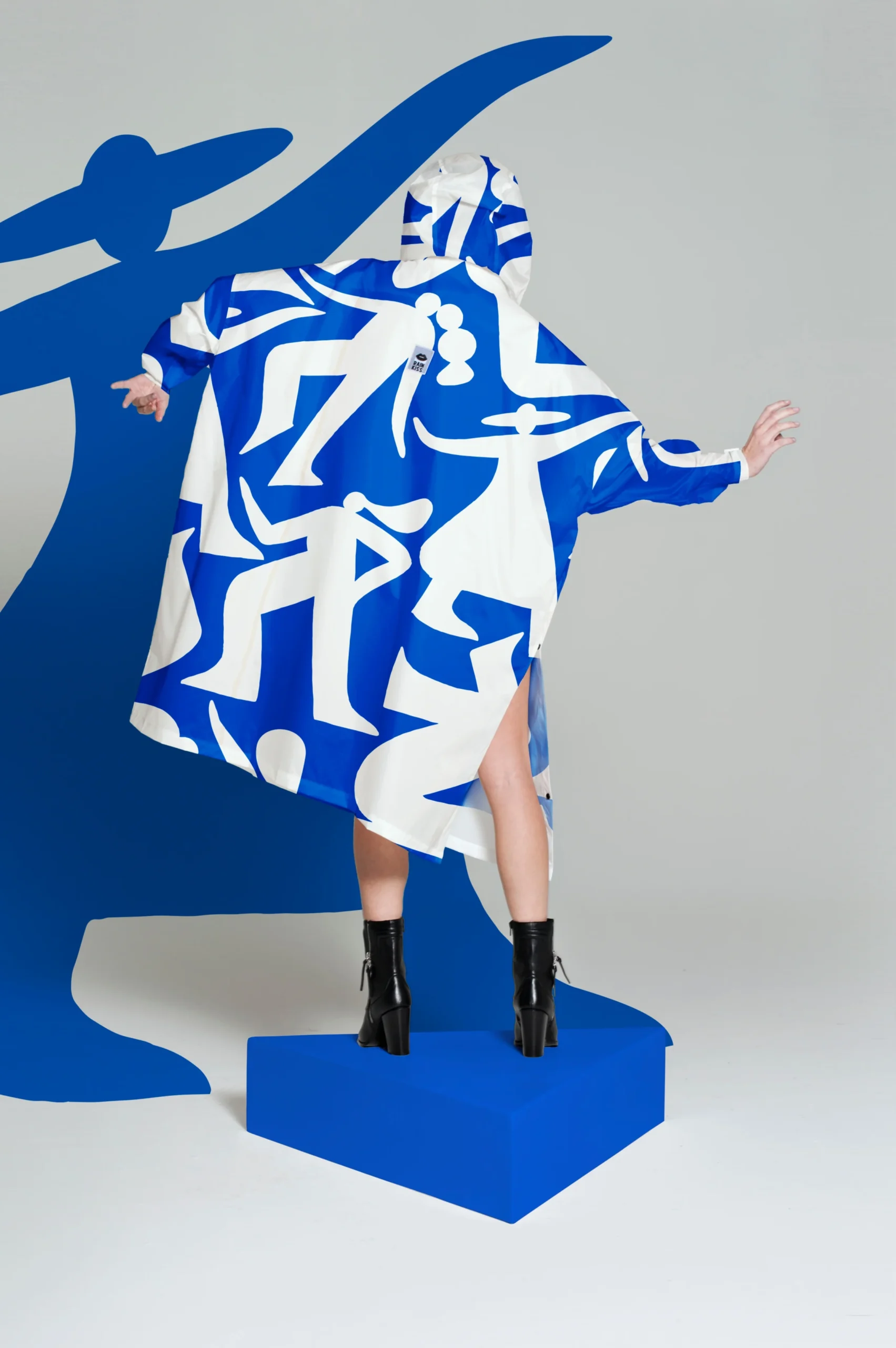A person models the Timo - Blue, a hooded garment with bold blue and white abstract designs inspired by Tallinn shopping. Styled for those seeking unique ideas, the look is finished with black heeled ankle boots on a blue platform. Gate21.ee