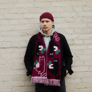 A man stands against a light brick wall, wearing a maroon beanie, light grey hoodie, black jacket, and the Cat - Organic scarf in maroon with cat and abstract designs. His hands are tucked into his jacket pockets. Gate21.ee