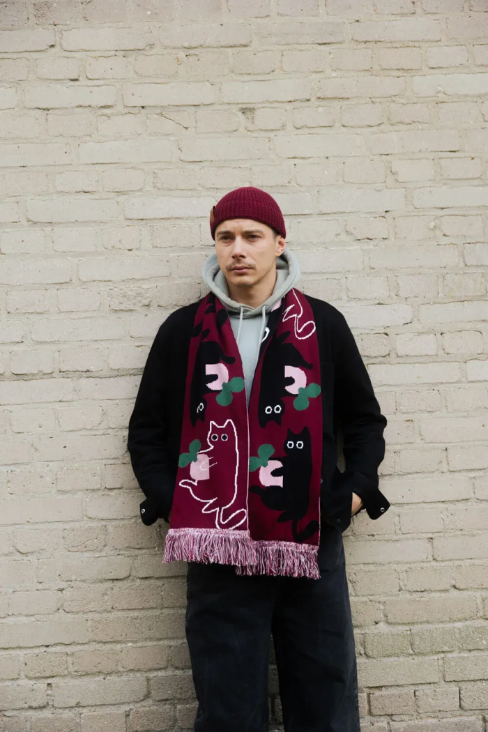A man stands against a light brick wall, wearing a maroon beanie, light grey hoodie, black jacket, and the Cat - Organic scarf in maroon with cat and abstract designs. His hands are tucked into his jacket pockets. Gate21.ee