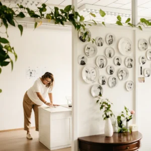A person stands at a white desk in a bright room, reviewing paperwork. The wall displays "The Glam Rocker - Big cup" with black-and-white Estonian designs, adding a touch of Tallinn gift shop charm alongside green plants and flowers. Gate21.ee