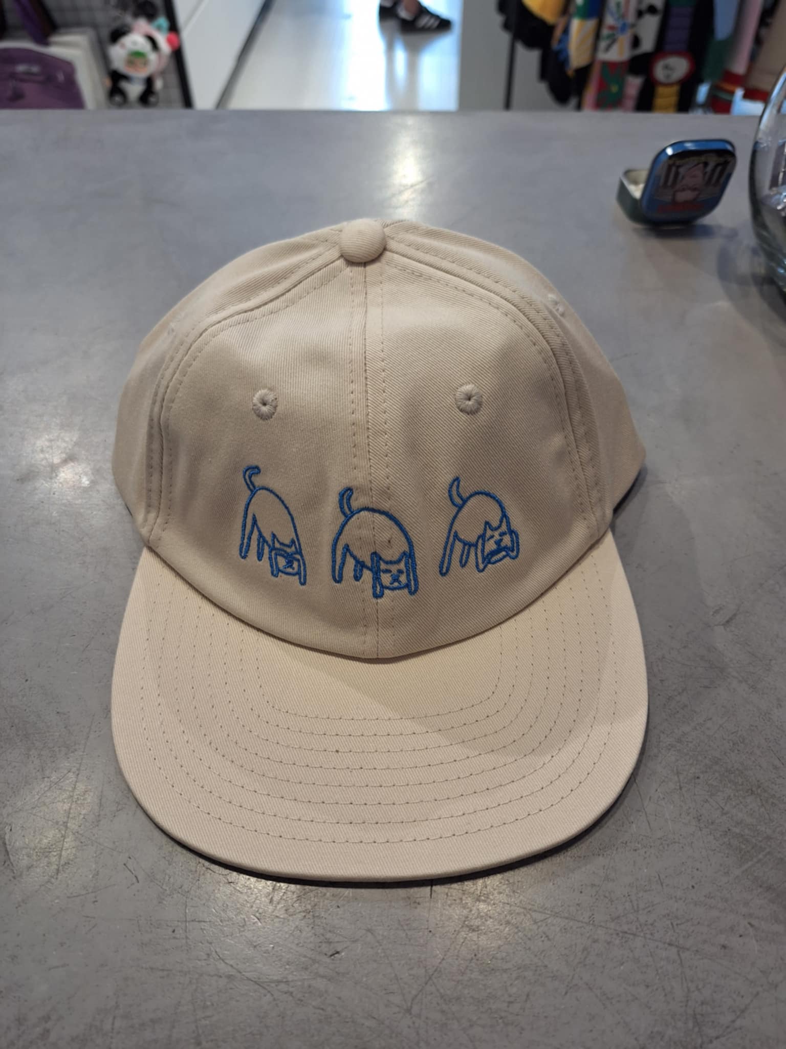 The Blonded Strapback hat - Off white sits on a grey countertop, featuring three simple blue line dog drawings across the front—an Estonian design ideal for anyone looking for unique gift shop ideas. Gate21.ee