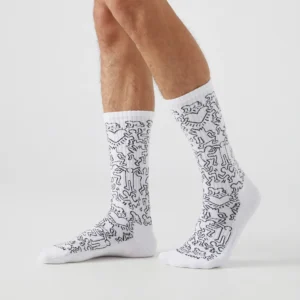 A person stands on a white surface, wearing Keith Haring Community - Athletic crew socks with black abstract line drawings. The mid-calf socks, shown on visible lower legs, make a unique addition to any gift shop. Gate21.ee