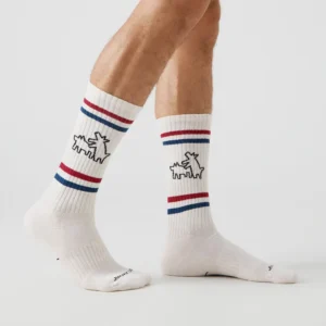 Someone wears Keith Haring Two Dogs - Athletic white crew socks with red and blue stripes and a stitched dog outline on each side, highlighting unique Estonian design. The legs are shown against a plain white background. Gate21.ee