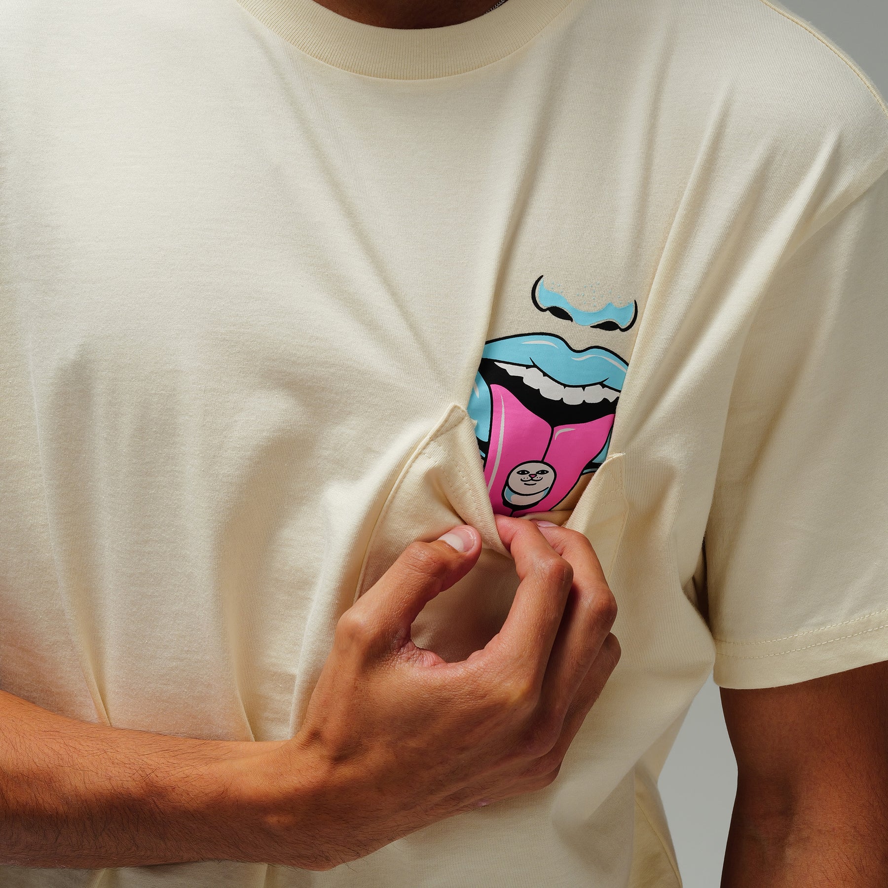 A person in the Take Your Meds Pocket Tee - Natural pulls the shirt to reveal a colourful cartoon face with a blue mouth and pink tongue on the chest pocket. Gate21.ee