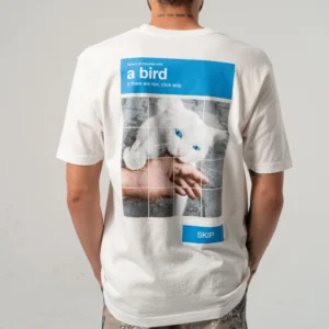 A person wears the A Bird Tee - Bone, featuring a blue-eyed white cat and a 'select all squares with a bird' CAPTCHA on the back. They’re facing away from the camera—perfect for concept shops or gift shops. Gate21.ee