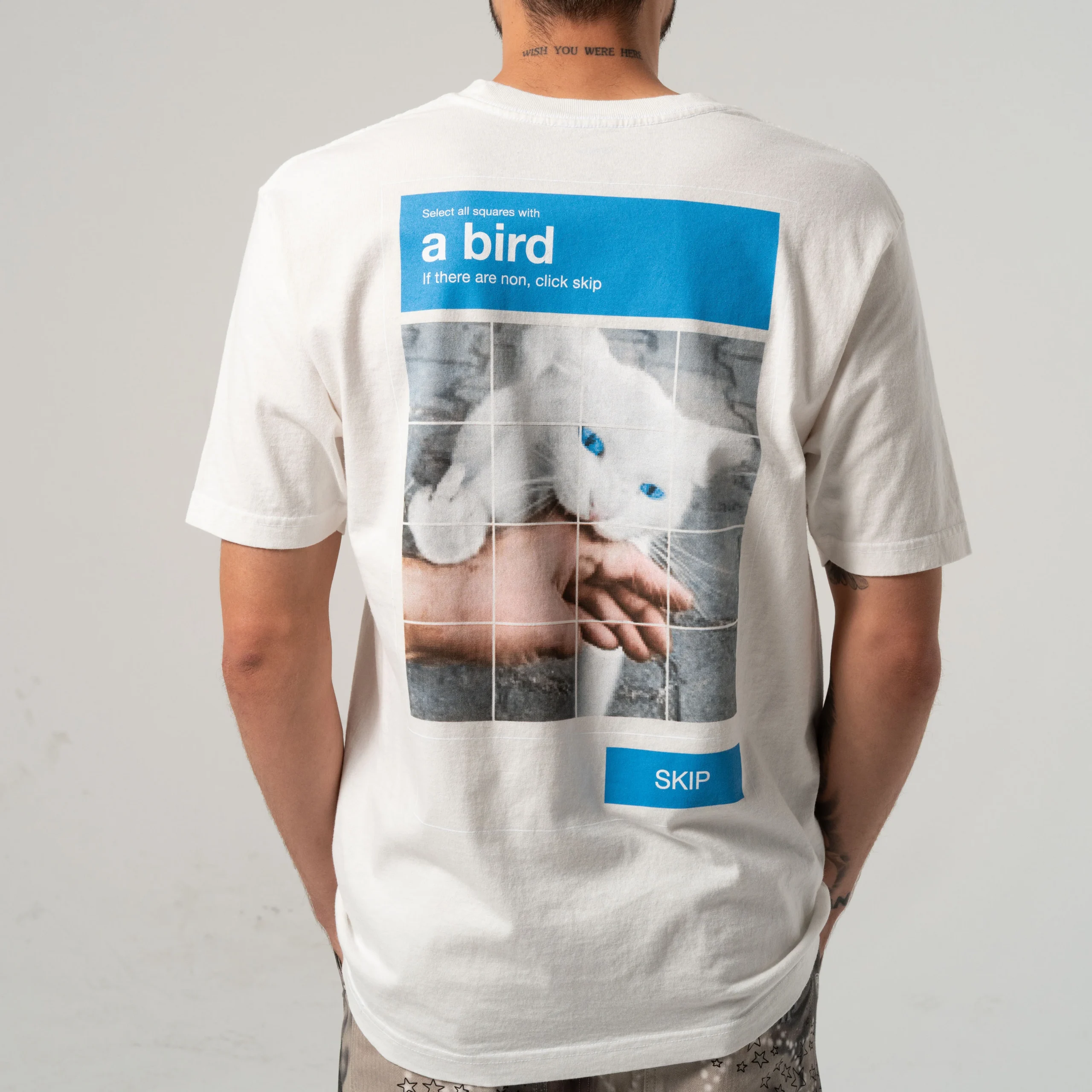 A person wears the A Bird Tee - Bone, featuring a blue-eyed white cat and a 'select all squares with a bird' CAPTCHA on the back. They’re facing away from the camera—perfect for concept shops or gift shops. Gate21.ee
