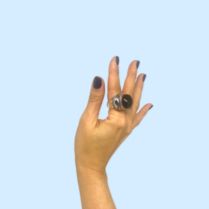 A hand with dark nails poses against a light blue background, wearing the Miss Nobody transparent/black ring—a large, oval statement piece inspired by Estonian design, perfect for Tallinn shopping or as a unique present. Gate21.ee