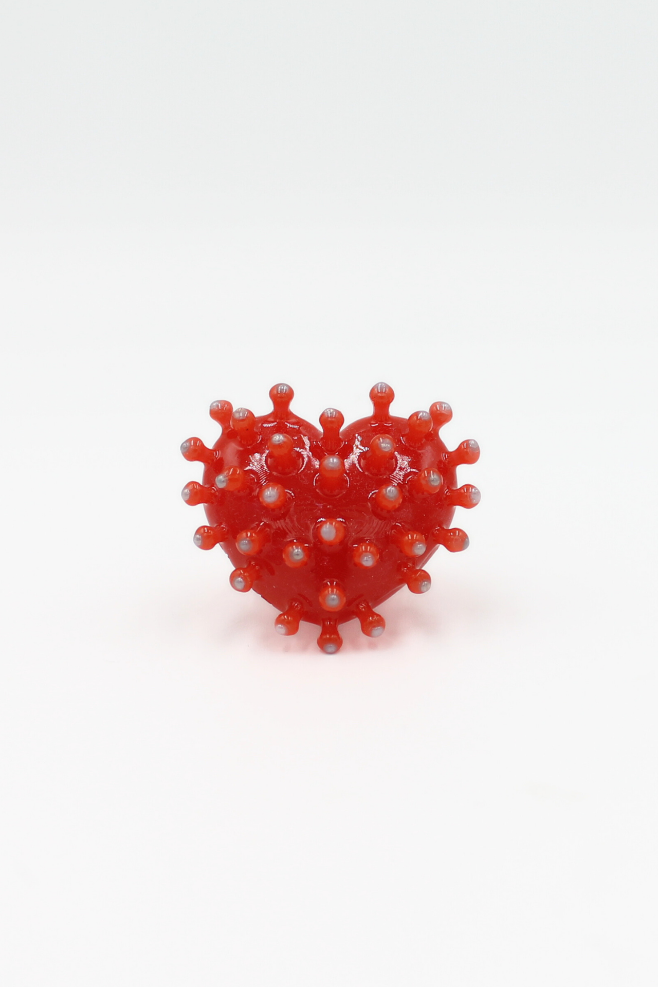 A red, heart-shaped object covered with round protuberances—resembling a stylised model of a virus—is centred against a plain white background. The object is decorated with CACKO rings and is identified as the “Virus - Ring.”. Gate21.ee