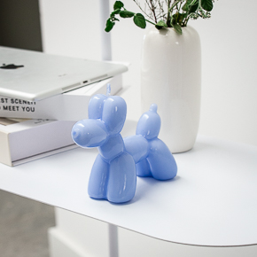 A light blue Balloon Dog Candle sits on a white table beside a curated stack of books and a white vase with green leafy branches, bringing the stylish charm of a modern concept shop to your décor. Gate21.ee