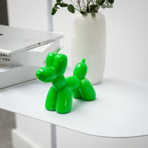 A Balloon Dog Candle in green sits on a white surface next to a white vase with leafy branches, offering unique decor inspiration. In the background, you'll spot a tablet and a closed book—evoking concept shop style. Gate21.ee