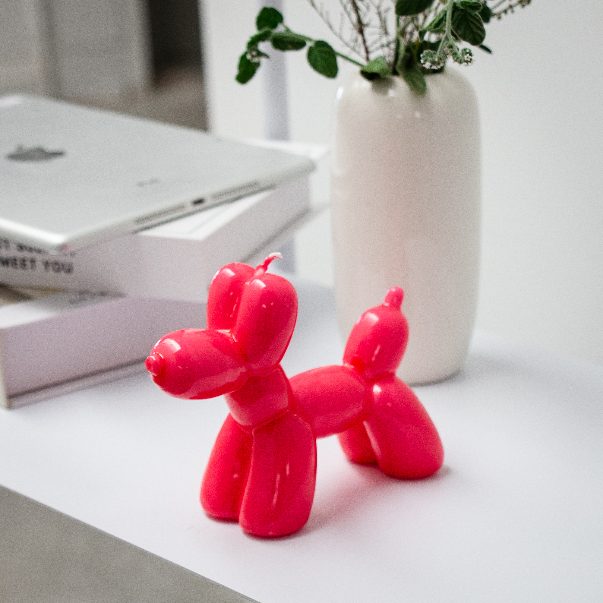 The Balloon Dog Candle - Magenta sits on a white surface beside a vase with green foliage and stacked books topped by a tablet—an ideal choice for anyone seeking unique finds from a curated gift shop. Gate21.ee