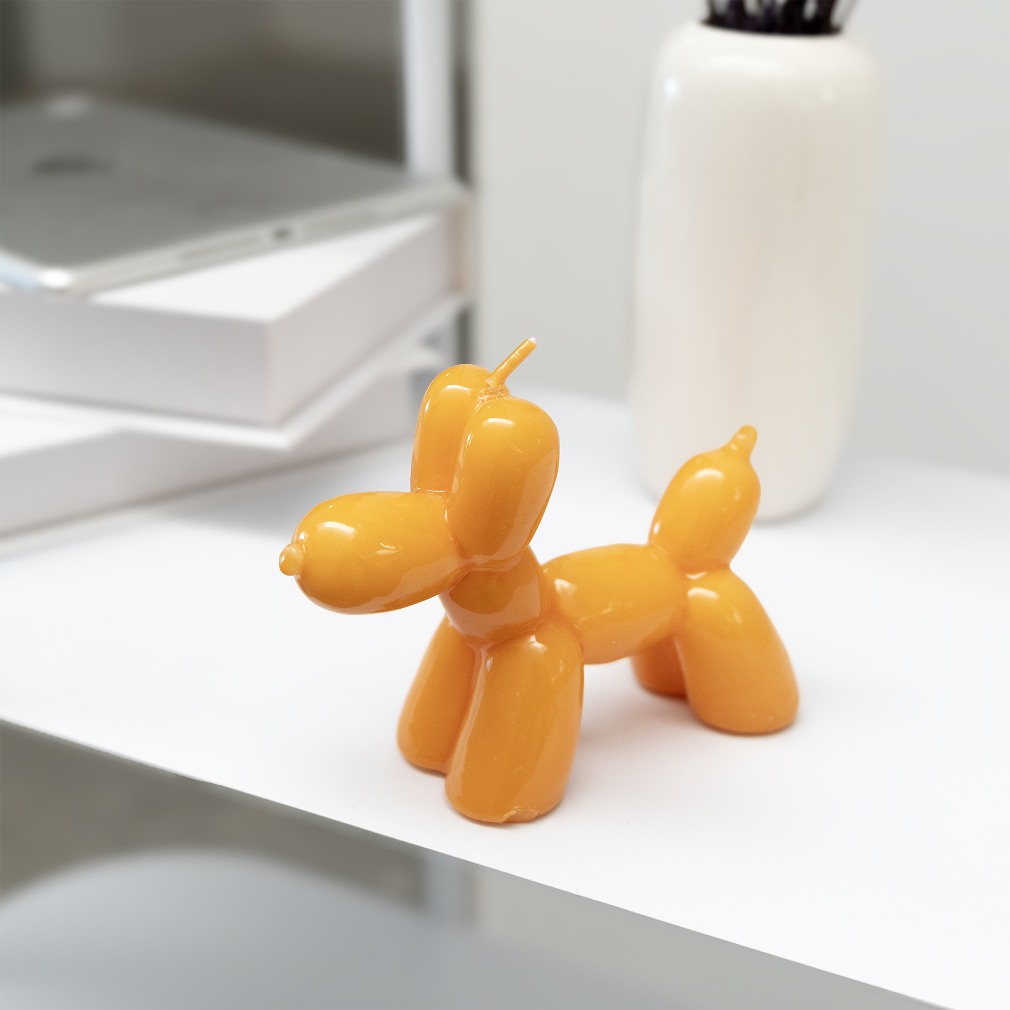 The Balloon Dog Candle - Orange sits on a white surface with a white vase and stacked books, creating striking inspiration for concept shop displays with its playful design and vibrant colour. Gate21.ee