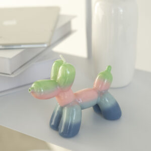 The Balloon Dog Candle - Sunrise, in pastel hues, rests on a white surface beside stacked books and a vase—ideal for curated concept shop displays or as a charming gift shop find. Gate21.ee