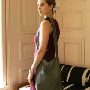 A woman with light skin and a white hair accessory stands indoors, wearing a sleeveless burgundy dress and carrying a Tinne+mia Feel Good Bag in Olive Night over her shoulder. She looks over her shoulder, with a black-and-white striped sofa in the background. Gate21.ee