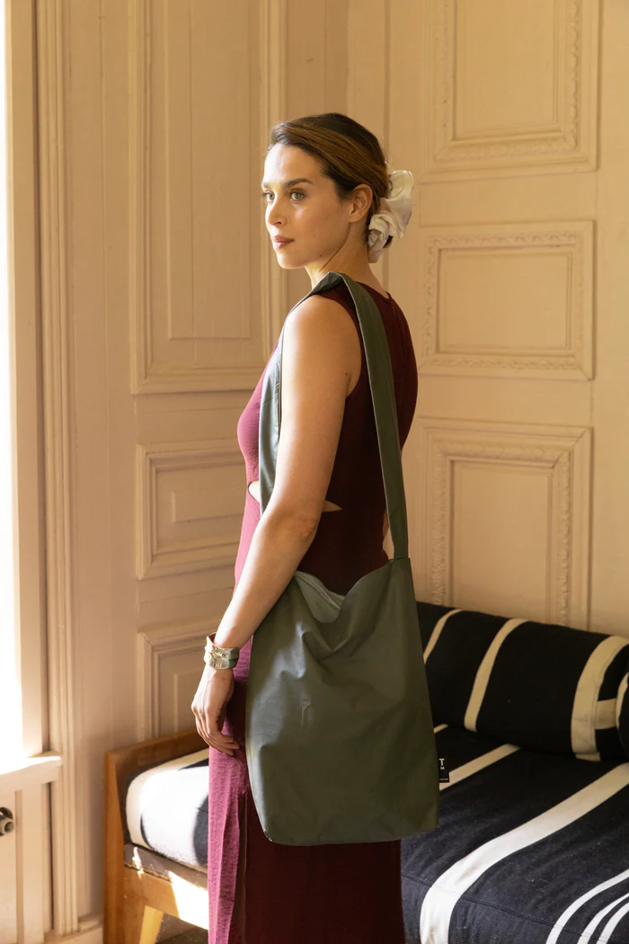 A woman with light skin and a white hair accessory stands indoors, wearing a sleeveless burgundy dress and carrying a Tinne+mia Feel Good Bag in Olive Night over her shoulder. She looks over her shoulder, with a black-and-white striped sofa in the background. Gate21.ee