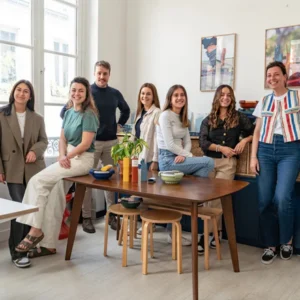 Seven people smile together in a bright, modern kitchen. With plants and dishes on the wooden table, the group use the Bini Cutlery Kit - Yellow as they sit and stand casually for a cheerful gathering. Gate21.ee