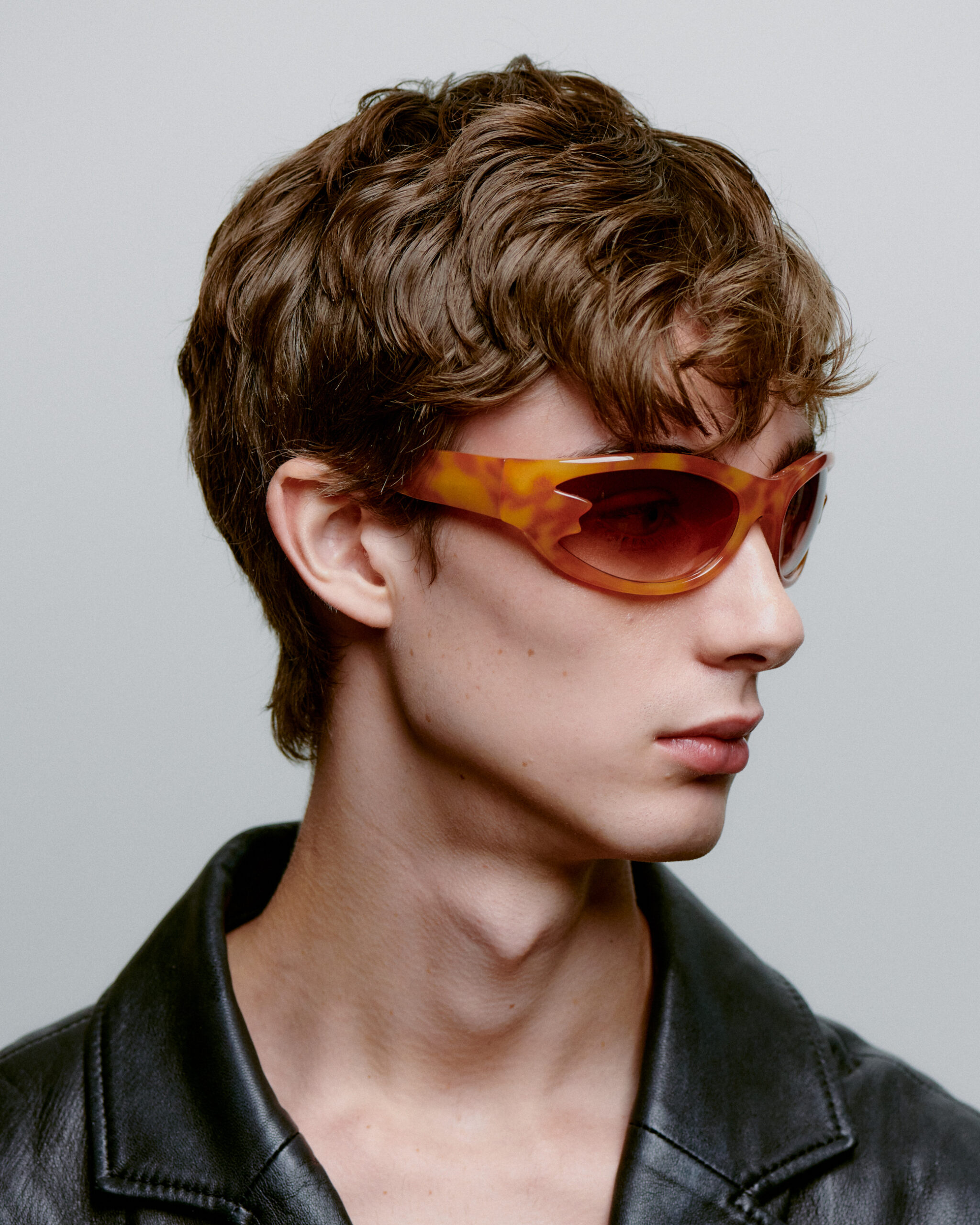 Wearing BLAST - Burnt Amber sunglasses, a person with short, wavy brown hair and a black leather jacket channels concept store style, gazing right against a plain grey background. Gate21.ee