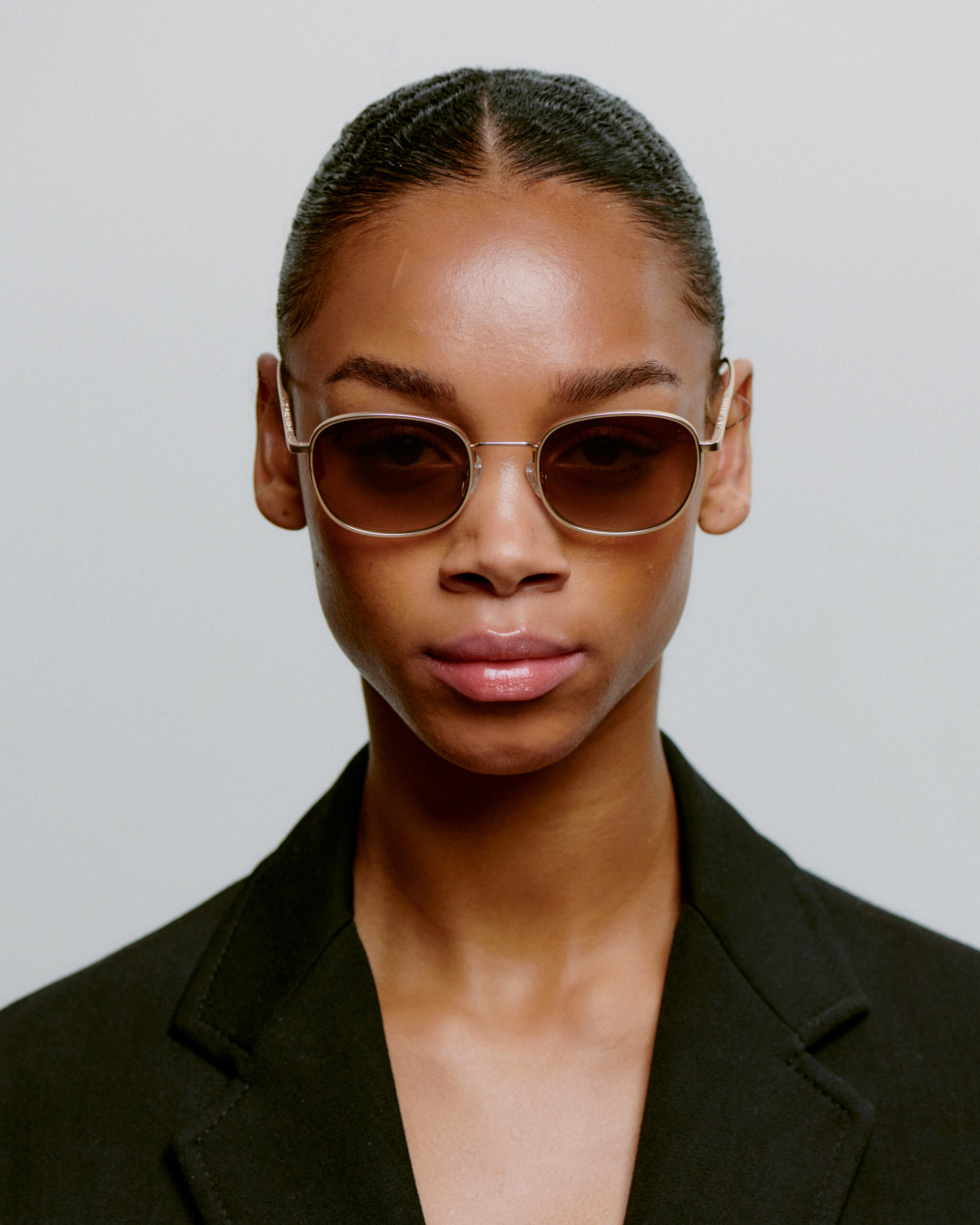 A person with slicked-back hair wears HELLO - Matte Gold sunglasses and a black blazer, facing the camera against a plain light background—a look inspired by Tallinn's curated shopping and concept store style. Gate21.ee