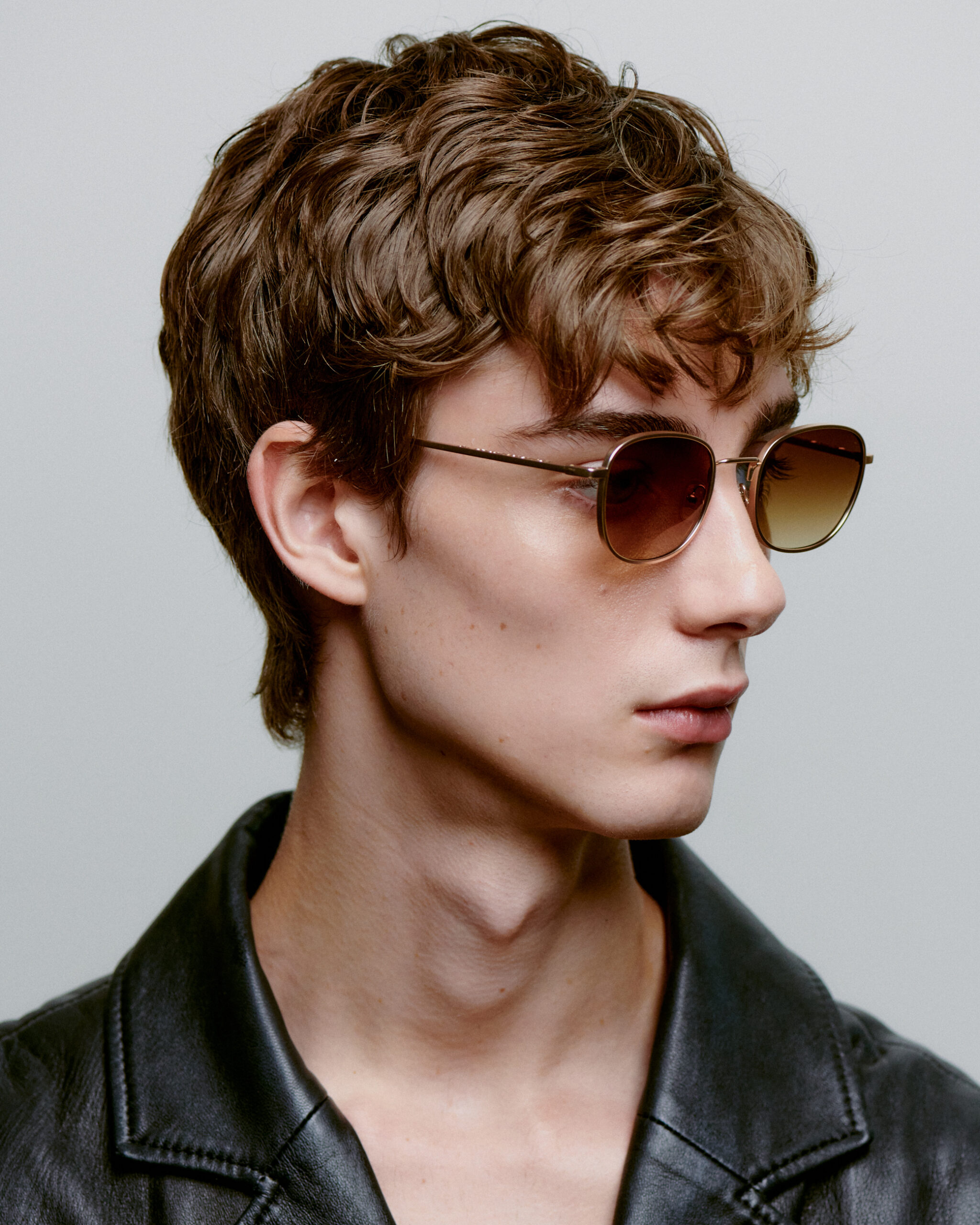 A person with short, wavy brown hair is wearing HELLO - Matte Gold sunglasses and a black leather jacket, looking slightly to the side against a plain background. Gate21.ee