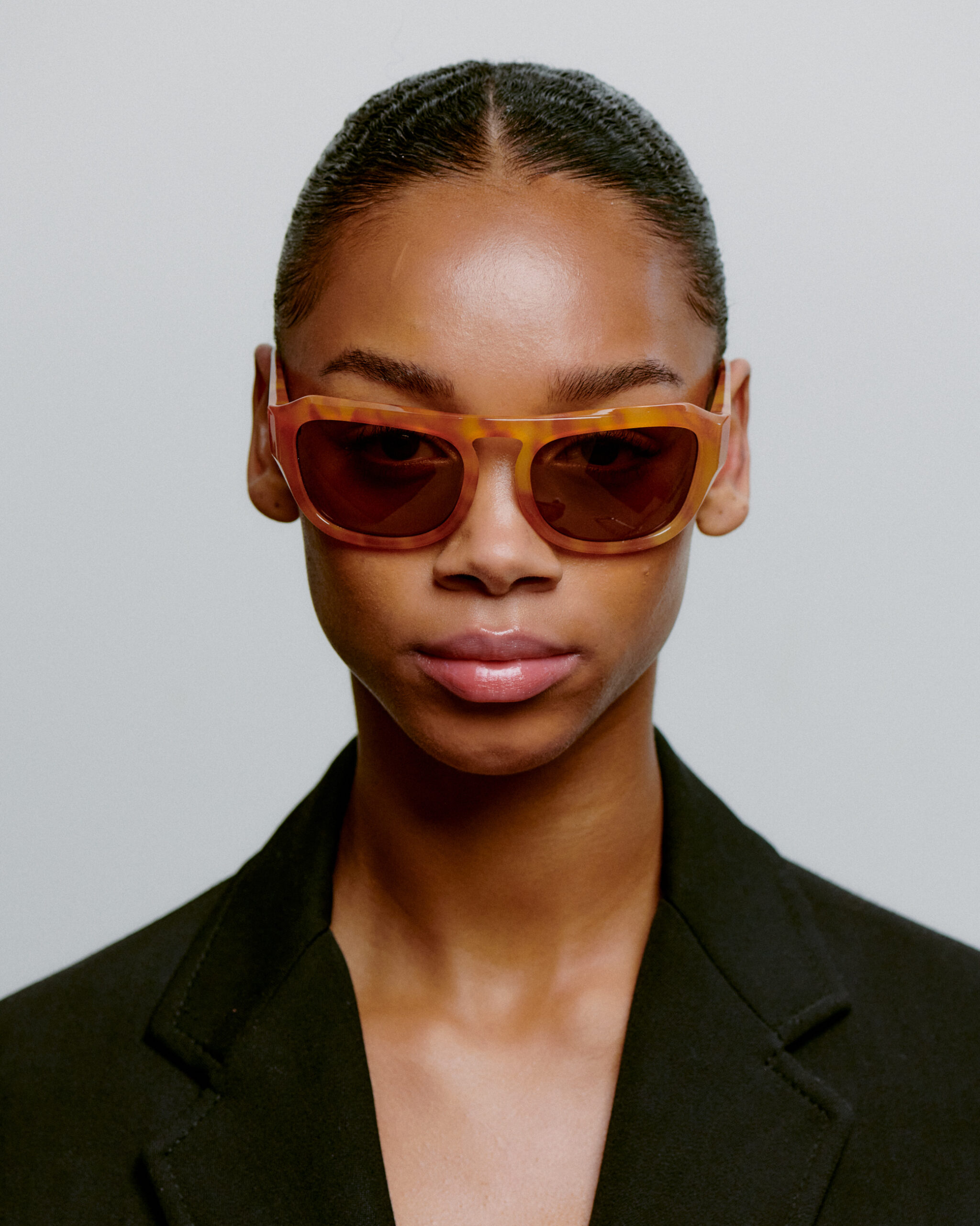 Wearing MAX - Burnt Amber sunglasses, a person with slicked-back hair and a black blazer faces the camera against a plain light background—exuding curated concept shop style. Gate21.ee