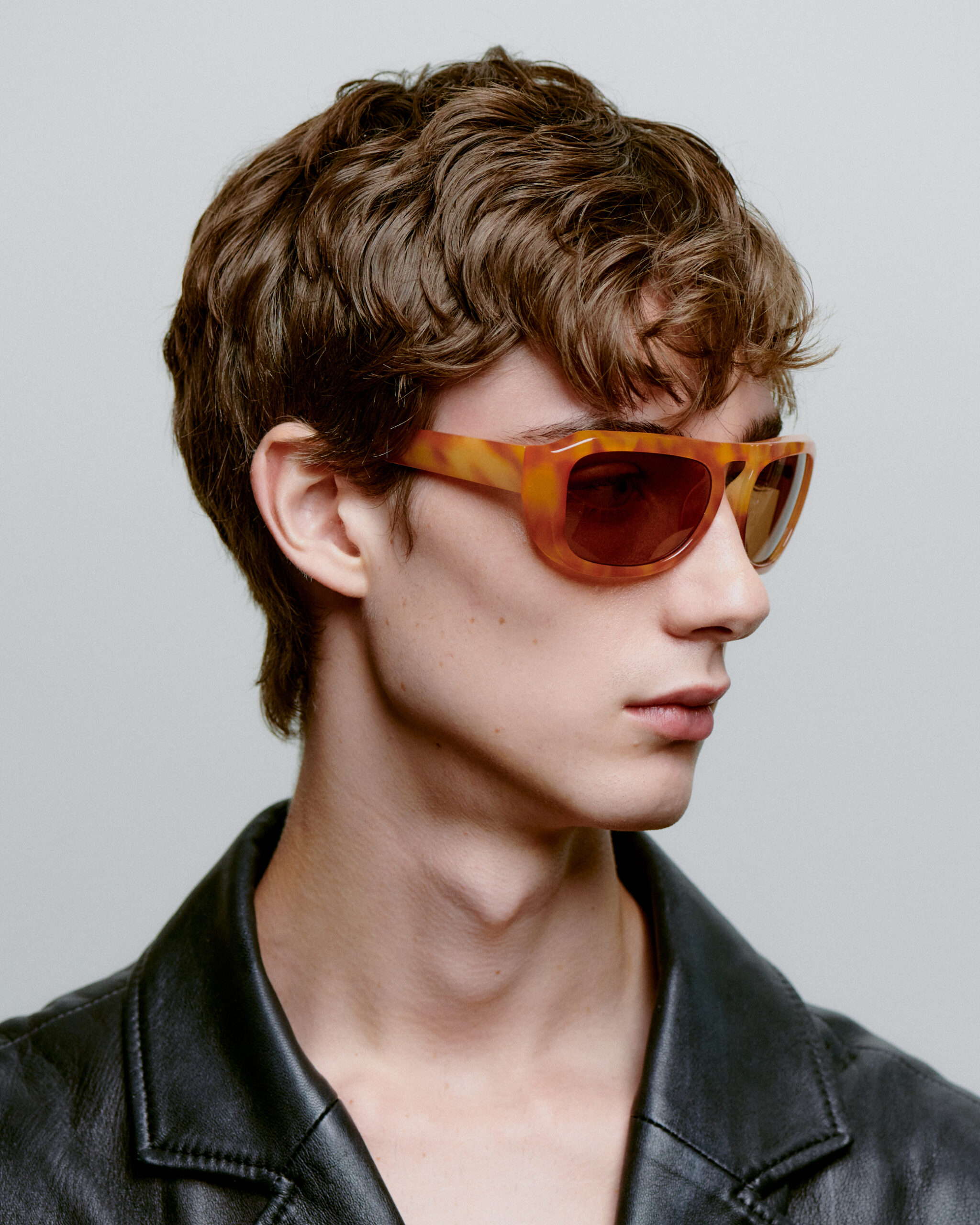 A person with wavy brown hair wears MAX - Burnt Amber sunglasses and a black leather jacket, glancing to the side against a plain light background, as if browsing creative ideas in a concept shop. Gate21.ee
