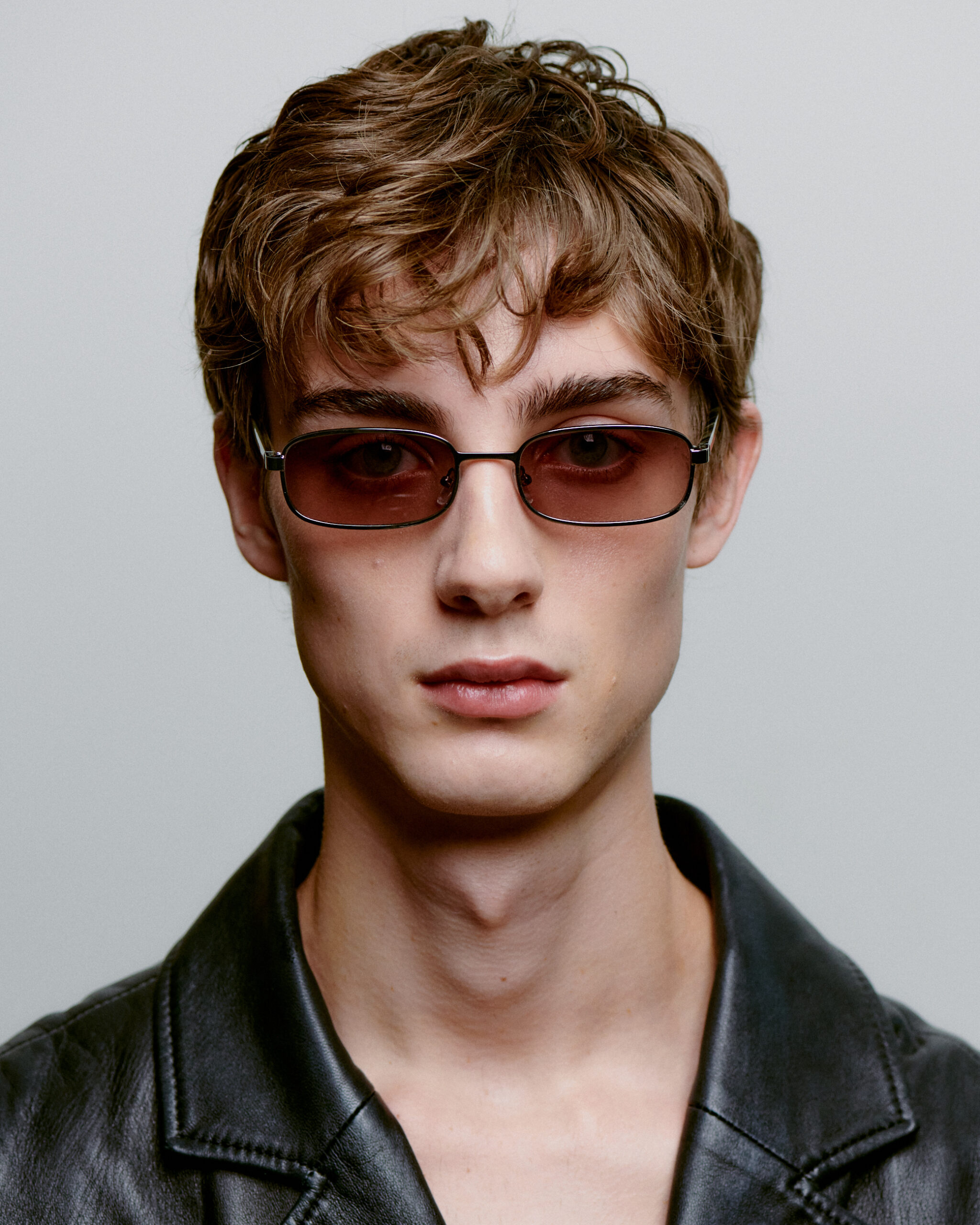 A young person with short, tousled brown hair wears NOAH - Silver Striped Horn rectangular sunglasses and a black leather jacket, facing the camera against a plain light background—perfectly embodying modern Estonian design. Gate21.ee