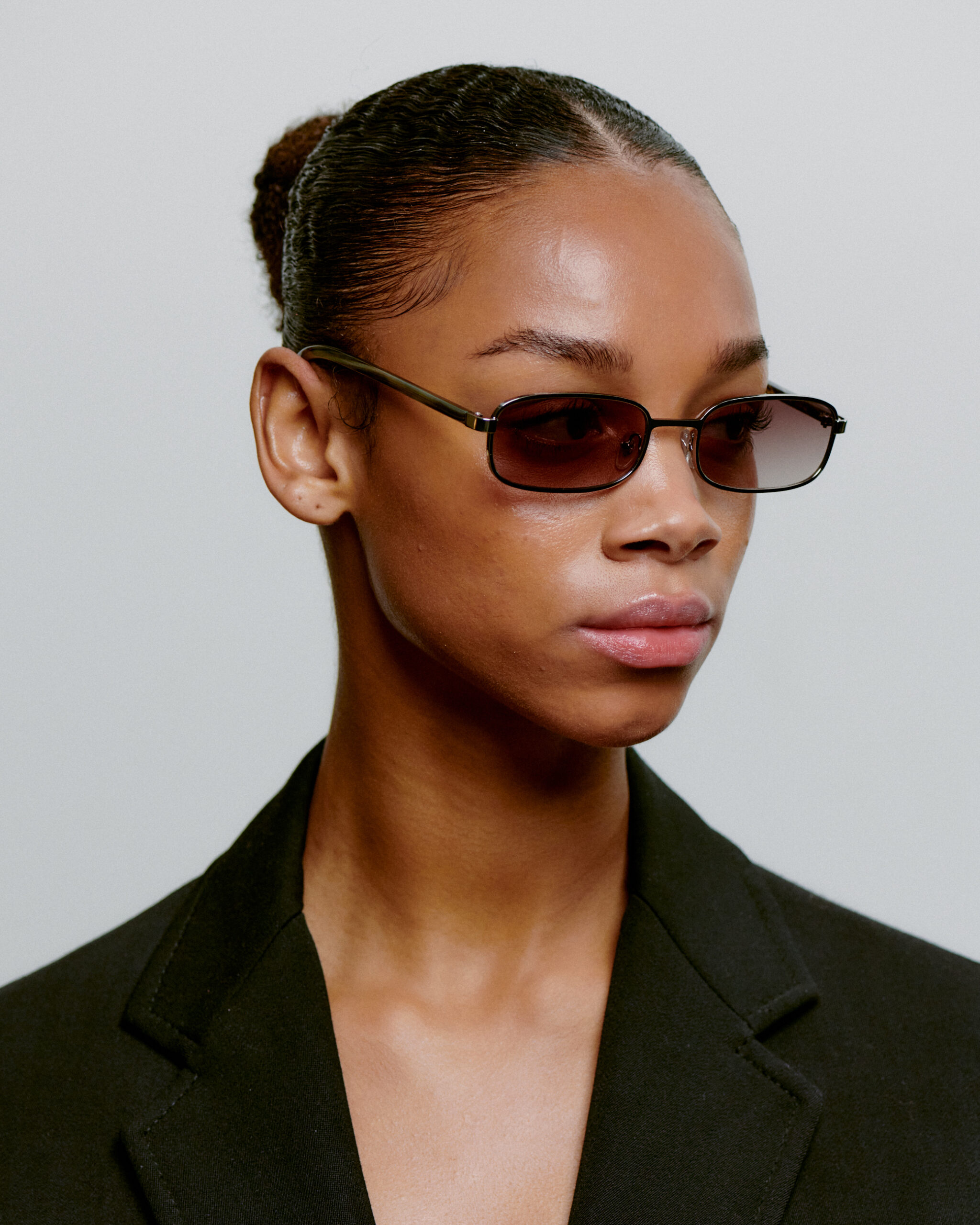 Wearing rectangular dark NOAH – Silver Striped Horn sunglasses and a black blazer, a person with slicked-back hair in a bun poses against a plain light grey background, reflecting the sleek minimalism of curated Estonian design. Gate21.ee