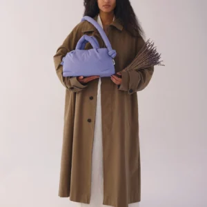 A person with long curly hair in a brown coat over a white dress holds dried lavender and the Mini Ona Soft Bag - Lilac—stylish inspiration from a Tallinn gift shop, set against a plain backdrop. Gate21.ee