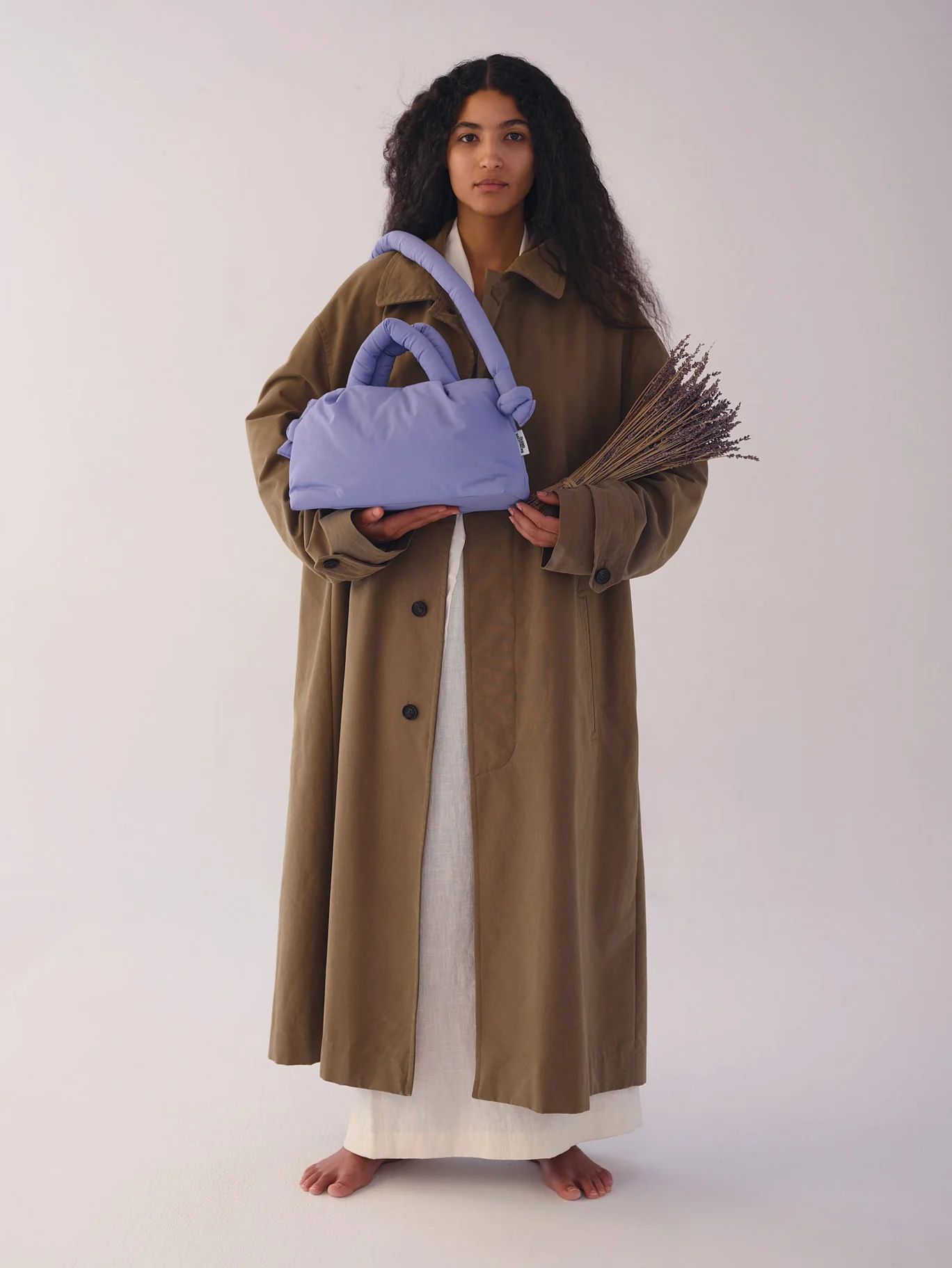 A person with long curly hair in a brown coat over a white dress holds dried lavender and the Mini Ona Soft Bag - Lilac—stylish inspiration from a Tallinn gift shop, set against a plain backdrop. Gate21.ee