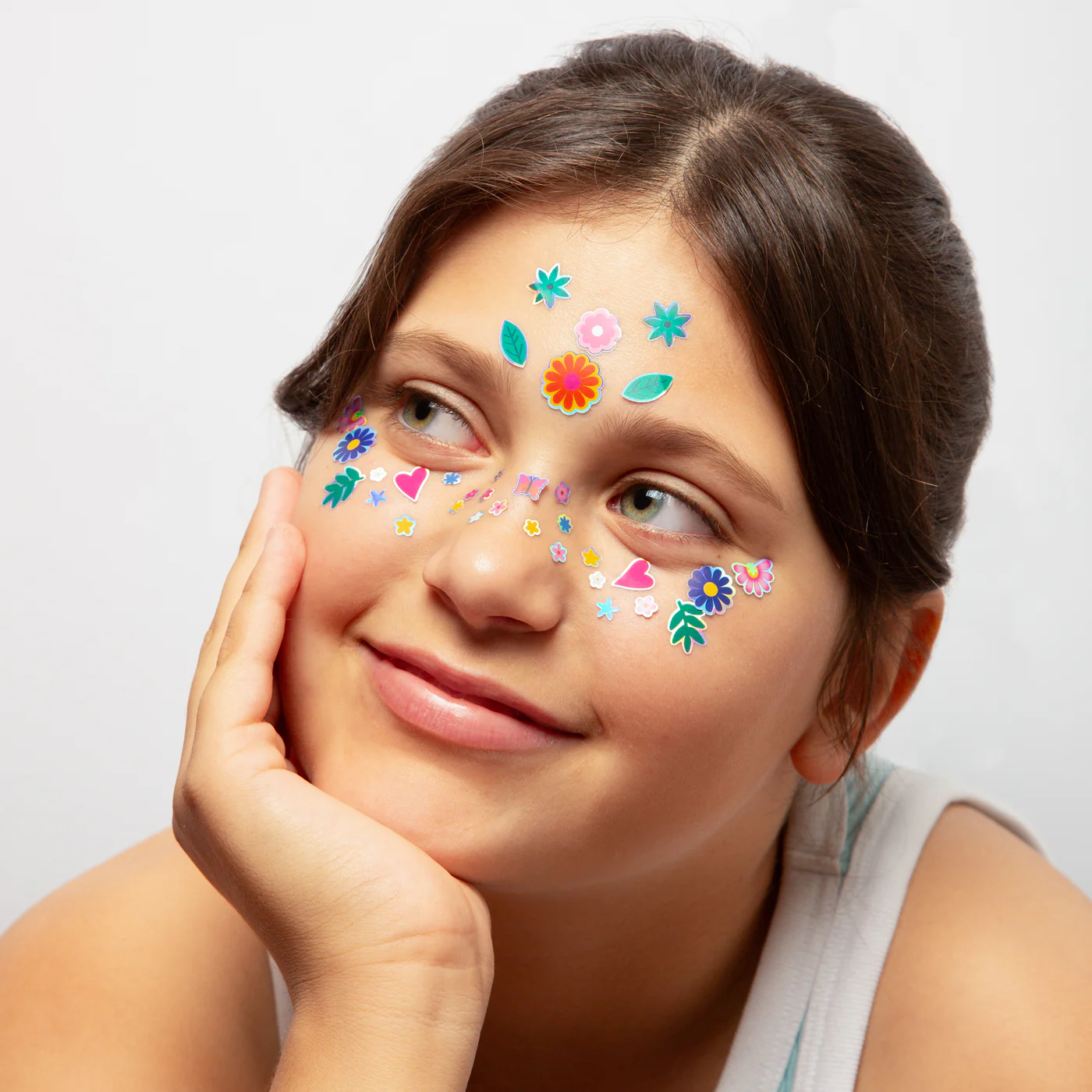 A girl with brown hair smiles, her chin resting on her hand. She wears a white vest and showcases Flower - Face Stickers on her forehead and cheeks—perfect inspiration for unique finds from a Tallinn gift shop. Gate21.ee